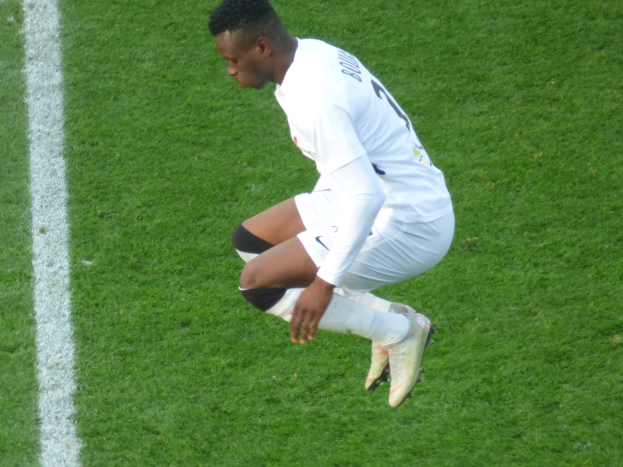 Berufsfußballspieler in weißer Uniform auf einem Fußballfeld kniend, Fußballschuhe tragend.