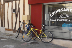 Zwei Fahrräder nebeneinander auf einem Gehweg vor einem Geschäft mit Glaswand geparkt, mit Text und einem Objekt links von den Fahrrädern.