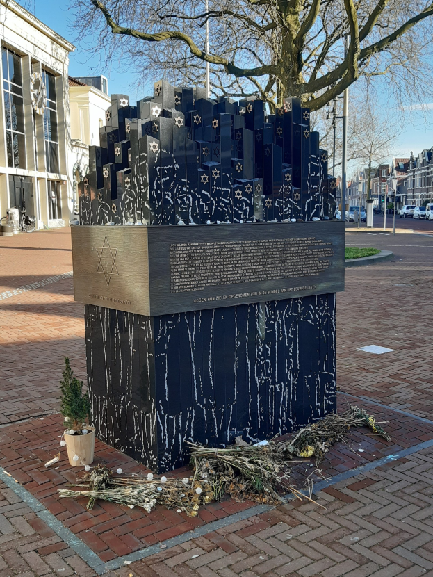 Denkmal auf einem Stadtplatz für Holocaust-Opfer mit einem Baum davor, einem Topfgewächs, verstreuten trockenen Blättern und Gebäuden, Fahrzeugen, Pfählen und Bäumen im Hintergrund.