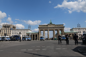 Gruppe von Polizisten vor dem Brandenburger Tor in Berlin, Deutschland, mit den Säulen des Tors, einer Statue, umgeben von Gebäuden, Fahrzeugen, Laternenmasten, Verkehrszeichen und bewölktem Himmel.