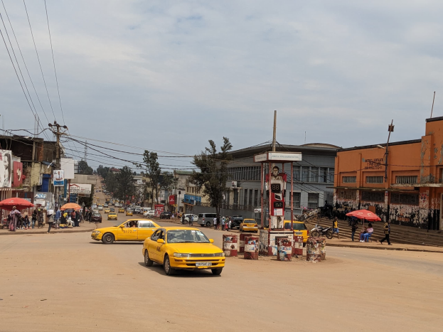 Ein gelbes Taxi fährt auf einer belebten Stadtstraße mit Gebäuden an den Seiten, während Fußgänger mit Regenschirmen auf dem Gehweg unter einem bewölkten Himmel gehen, mit Strommasten und Kabeln im Hintergrund.