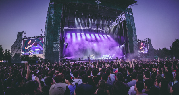 Eine große Menschenmenge auf einem Musikfestival mit einer Bühne, auf der Musiker spielen, beleuchtet von Lichtern und umgeben von Bildschirmen, vor einem Gebäude, Bäumen und einem klaren blauen Himmel.