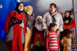 Eine Gruppe von Menschen, einige in traditioneller Kleidung und andere mit Kopftuch, steht zusammen vor einem Zelt mit einer Wand im Hintergrund und sieht aus wie Flüchtlinge.