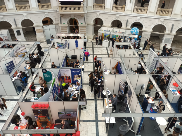 Eine große Gruppe von Menschen auf einer internationalen Fachmesse mit Ständen, Bannern, Tischen, Stühlen und Luftballons in einem Raum mit architektonischen Bögen, Säulen und einem Bildschirm im Hintergrund.