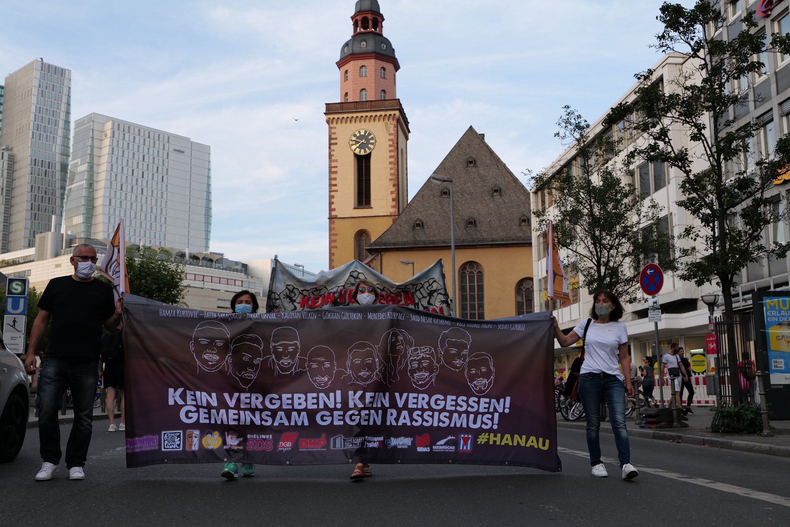 Eine Gruppe von Menschen in Masken, die eine Straße entlanggehen und ein Banner halten, mit einem geparkten Auto auf der linken Seite, Gebäuden, Bäumen, Schildern, Pfählen, einem Uhrenturm und einem klaren blauen Himmel im Hintergrund.