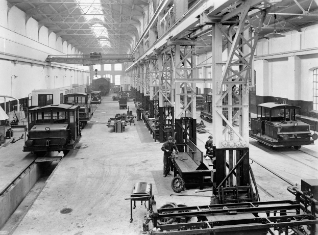 Schwarzes und weißes Foto einer Fabrikhalle mit mehreren trains unter Reparatur, Arbeitern, die sich darum kümmern, Metallpfosten, die das Dach stützen, Deckenlampen, die den Raum beleuchten, und Fenstern an den Wänden.