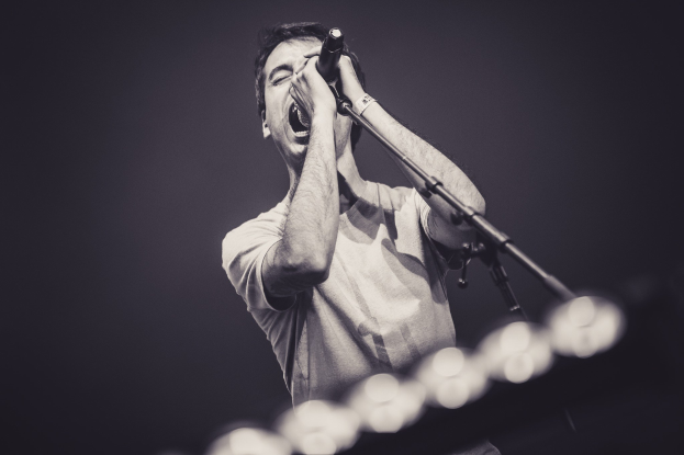 Ein Mann in schwarzem T-Shirt und Jeans singt leidenschaftlich in ein Mikrofon auf einer hell erleuchteten Bühne, seine Augen sind geschlossen und seine Haare sind stylisch durcheinander.