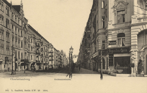Altes Schwarz-Wei├č-Foto einer Stadtstra├če in Charlottenburg, Deutschland, mit Geb├Ąuden, Stra├čenlaternen, L├Ąmpchen, Namensschildern, Kraftfahrzeugen, Passanten, Gittern und einem klaren Himmel.