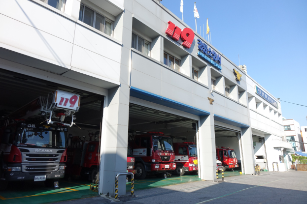 Feuerwache mit mehreren Feuerwehrautos vorne, mit Fenstern, Säulen, Namensschildern und Flaggen mit Stangen im Vordergrund vor einem Hintergrund aus Gebäuden und einem klaren blauen Himmel.