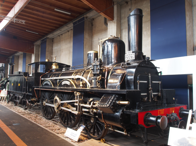 Alte Dampflokomotive in Museums-Umgebung mit umgebenen Eisenbahnschienen, Informationsschildern und anderen Ausstellungsstücken unter Deckenbeleuchtung.