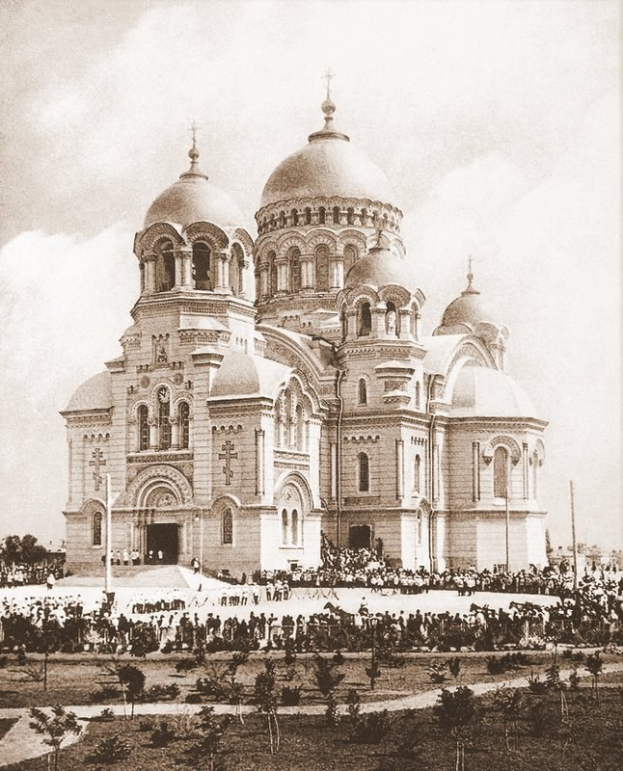 Schwarz-weiß-Fotografie der Erlöserkirche auf dem Bluteigen in St. Petersburg, Russland, mit Menschen, die sich um das prunkvolle Gebäude versammeln, Bäumen und Pfählen im Vordergrund und einem klaren Himmel im Hintergrund.