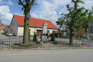 Ein kleines Dorf mit einer Kirche in der Mitte, umgeben von einem Metallzaun, Bäumen, Pflanzen, Gras, einem Schild, Fahrzeugen, Polen, Drähten und einem bewölkten Himmel; die Kirche ist die Kirche des Heiligen Grabes in St. Vitus.