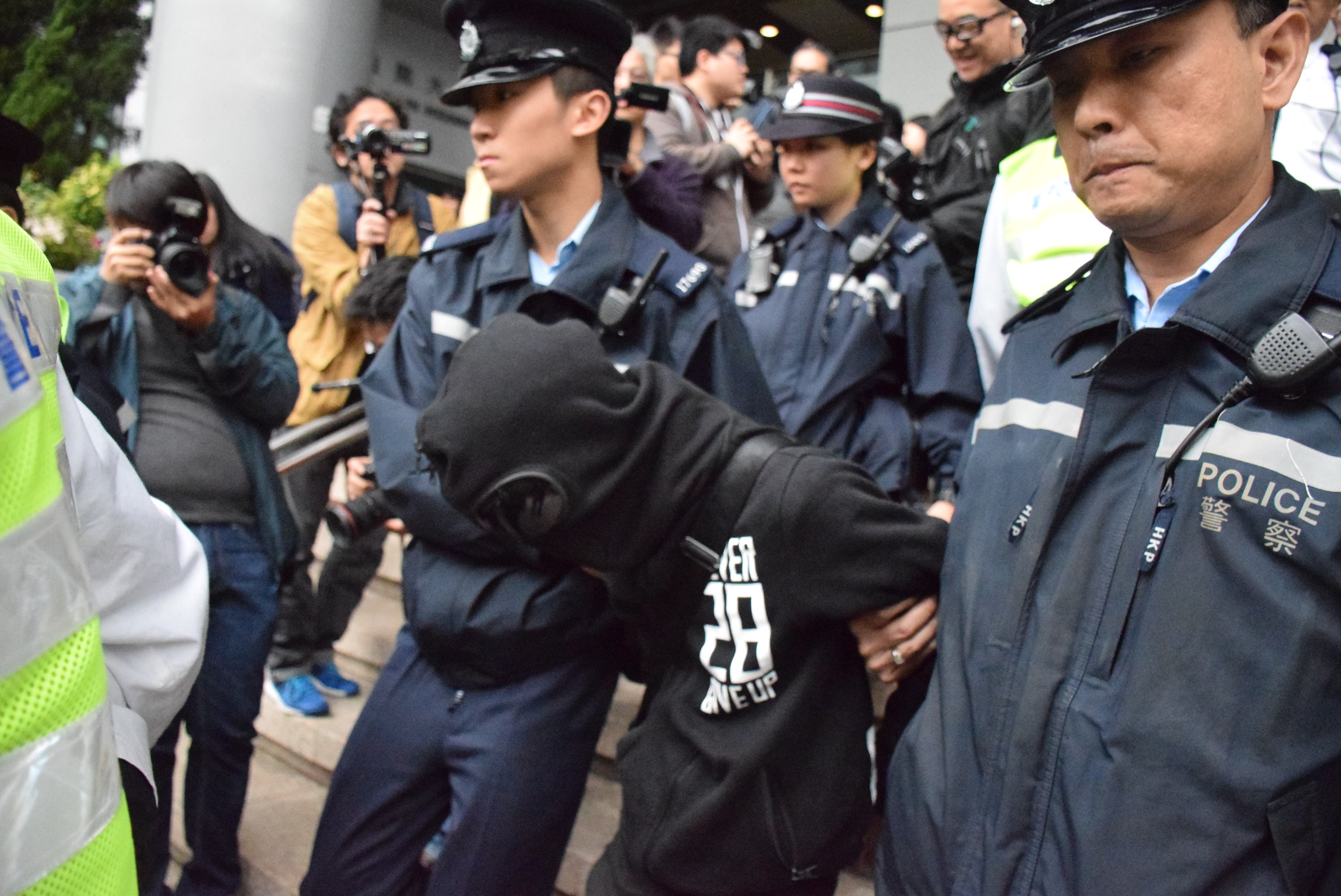 Eine Gruppe von Polizeibeamten steht vor einer Menschenmenge, einige tragen Mützen und halten Kameras, im Hintergrund ein Gebäude mit Säulen und Lampen und links Bäume.