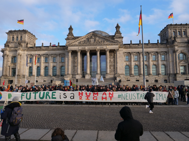 Gruppe von Menschen vor dem Reichstagsgebäude in Berlin mit einer Fahne mit der Aufschrift "Zukunft ist ein Mensch Neustar ima" und umgeben von Fahnenmasten.