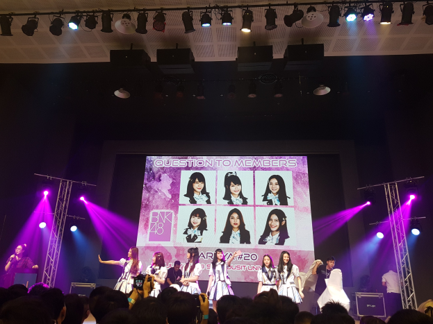 Eine Gruppe von Menschen, die auf einer hell beleuchteten Bühne performen, wobei einer ein Mikrofon hält und ein anderer ein Instrument spielt, während die Menge das Ereignis aufzeichnet; ein großer Bildschirm im Hintergrund zeigt Bilder der K-Pop-Gruppe BK48.