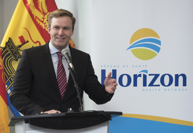 Ein Mann in einem schwarzen Blazer, weißem Hemd und roter Krawatte steht an einem Podium mit einem Mikrofon und einem Banner im Hintergrund.