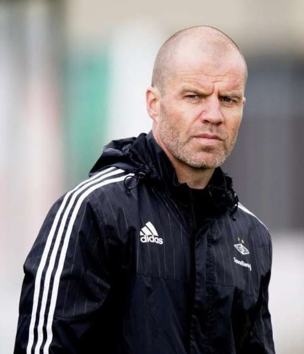 Ein kahlköpfiger Trainer in einer schwarzen Jacke steht auf einem Fußballfeld, trägt eine marineblaue Jacke mit weißen Streifen und hat die Hände vor der Brust verschränkt.