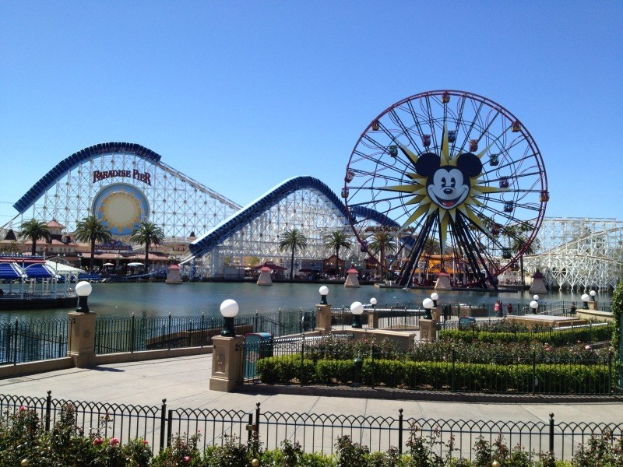 Luftaufnahme von Disney California Adventure mit seinem Riesenrad und Achterbahn, umgeben von Grünflächen und Laternenmasten, mit Wasser im Vordergrund und einem klaren blauen Himmel.