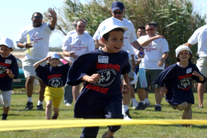 Kinder laufen auf einem Rasenfeld mit einem gelben Band im Vordergrund, einige tragen Kappen und Schutzbrillen, andere halten Gegenstände, vor einem Hintergrund aus Bäumen und einem klaren blauen Himmel.