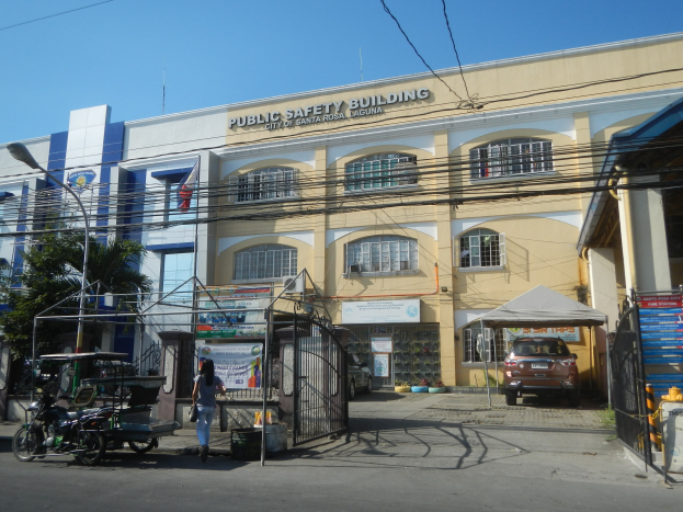 Gebäude für öffentliche Sicherheit mit umgebender Infrastruktur, einschließlich Bäumen, Straßenmöbeln, Fahrzeugen, Fußgängern und einem klaren Himmel.