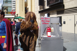 Eine Gruppe von Menschen, einige in Kostümen, versammelte sich vor Gebäuden mit sichtbarer Beschriftung, einer Box mit Schrift und einem Feuerlöscher.