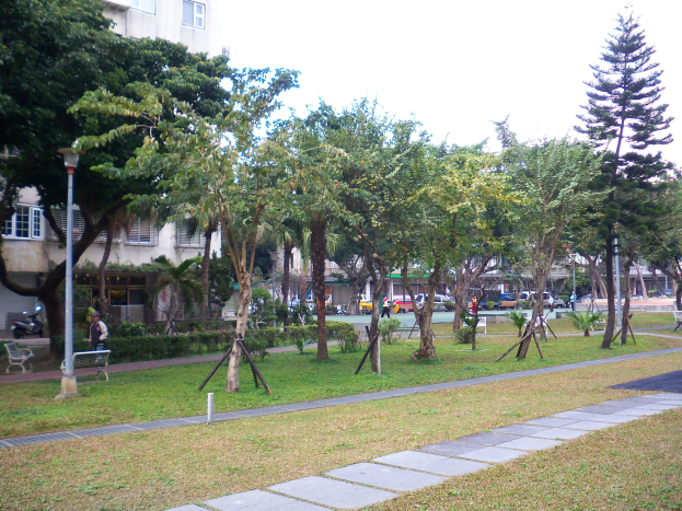 Ein Park mit grünem Gras, Bäumen und einem Weg mit Bänken und Laternen, der von Menschen, Fahrzeugen, Gebäuden und einem klaren blauen Himmel gesäumt ist.