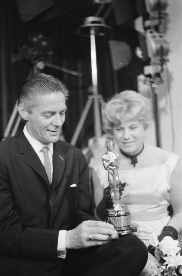 Ein Schwarz-Wei├č-Bild eines formell gekleideten Mannes, der eine Oscar-Statue hält und eine Frau, die einen Blumenstrau├č hält, nebeneinander sitzend mit beleuchtetem Hintergrund.