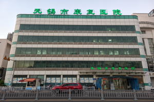 Ein großes Gebäude mit vielen Fenstern, das als chinesisches Krankenhaus bezeichnet ist, umgeben von einem Metallzaun, mit Fahrzeugen auf der Straße, Menschen auf dem Gehweg, einem Schild, Pflanzen und einem bewölkten Himmel.