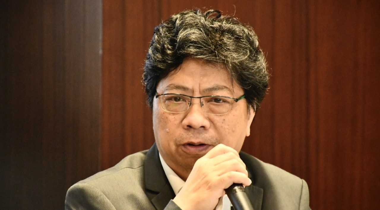 Ein Mann im Anzug mit einem Mikrofon, der lebhaft vor einem Holz-Hintergrund spricht.