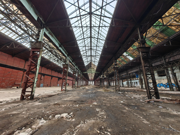 Verlassenes Gebäude mit verstreuter Trümmern, Metallstangen, einem beschädigten Dach mit Glasabschnitten, graffiti-bedeckten Wänden und zerbrochenen Fenstern.