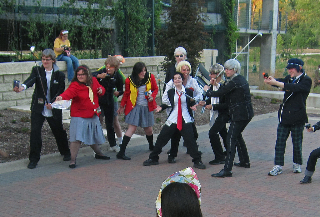Eine Gruppe von Menschen steht in der Mitte, einige halten Hockeystöcke, eine Frau auf der linken Seite hält einen Fächer; im Hintergrund sitzt eine Frau auf einer Wand mit Bäumen, einem Gebäude und Gras und einem Weg am Boden.