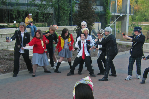 Eine Gruppe von Menschen steht in der Mitte, einige halten Hockeystöcke, eine Frau auf der linken Seite hält einen Fächer; im Hintergrund sitzt eine Frau auf einer Wand mit Bäumen, einem Gebäude und Gras und einem Weg am Boden.
