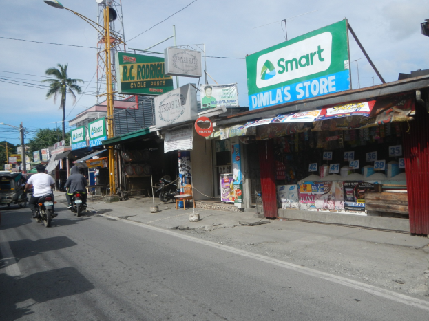 Eine belebte Straße mit Motorradfahrern, Autos, Gebäuden, Schildern, Strommästen, Kabeln, Straßenlampen, Bäumen und einem bewölktem Himmel.