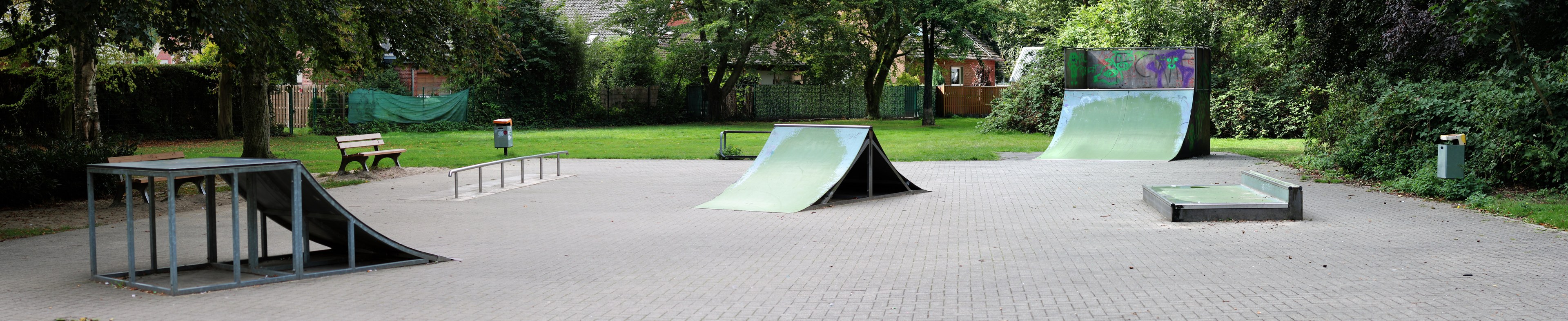Ein Park mit einer zentralen Skateboard-Rampe, umgeben von Bänken, Geländern, Gras, Pflanzen, Bäumen und Gebäuden im Hintergrund.