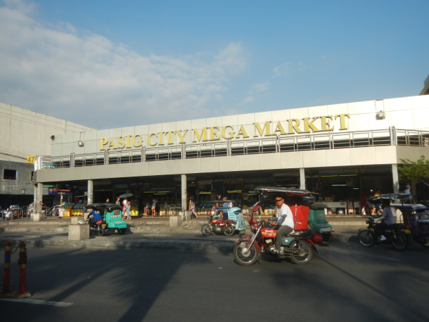 Eine belebte Straßenszene am Pasig City Mega Market mit Fahrzeugen und Fußgängern, die ein Gebäude mit sichtbarem Text im Hintergrund unter einem bewölkten Himmel zeigen.