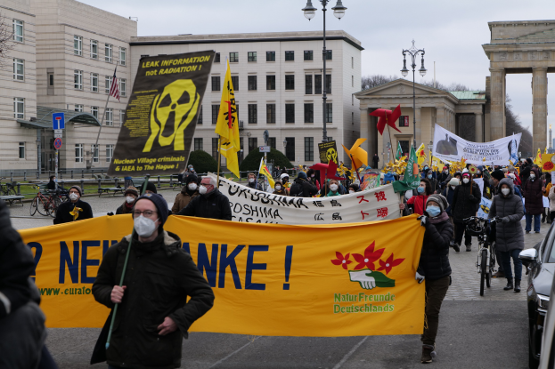 Große Gruppe von Menschen bei einer Demonstration gegen Atomkraft in Deutschland, mit Schildern und Fahnen, begleitet von Fahrzeugen und Gebäuden im Hintergrund.