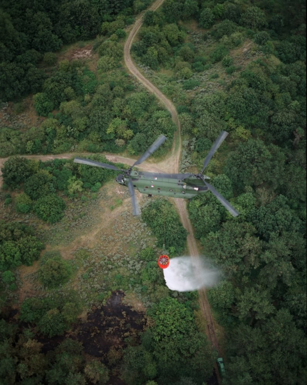 Ein Hubschrauber, der über einem dichten grünen Wald mit einer Straße im Hintergrund fliegt, und Text am unteren Bildrand.