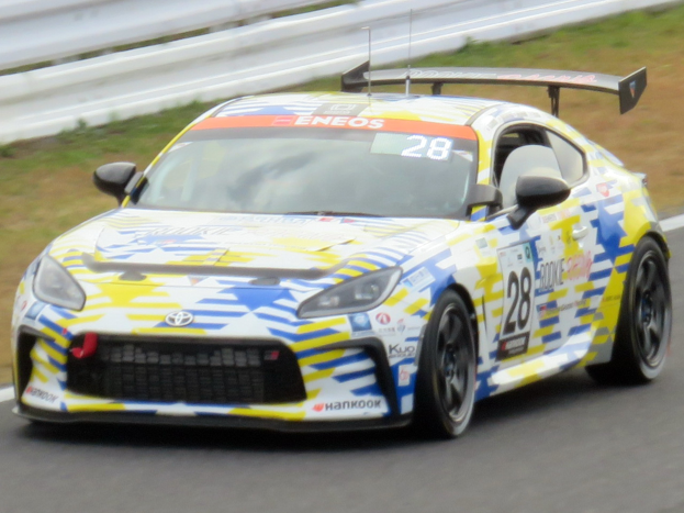 Ein weißer Toyota Supra GT3-Rennwagen mit Sponsorentext und Nummern, der auf einer Rennstrecke fährt, mit einer Begrenzung und Gras im Hintergrund.
