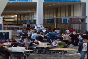 Ein belebter Flughafenvorplatz mit wartenden Menschen, einige stehen und andere arbeiten, Richtungsschilder an der rechten Wand und darüber sowie von der Decke hängende Lampen.