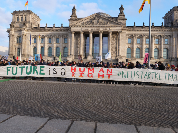 Eine Gruppe von Menschen hält ein Transparent mit der Aufschrift "Zukunft ist ein Mensch" vor dem Reichstaggebäude in Berlin, Deutschland, mit sichtbaren architektonischen Details und Flaggen im Hintergrund bei bewölktem Himmel.