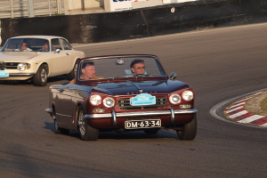 Zwei Rennautos auf einer Rennstrecke mit Zuschauern, Gras auf der rechten Seite und ein Zaun im Hintergrund mit Texttafeln.