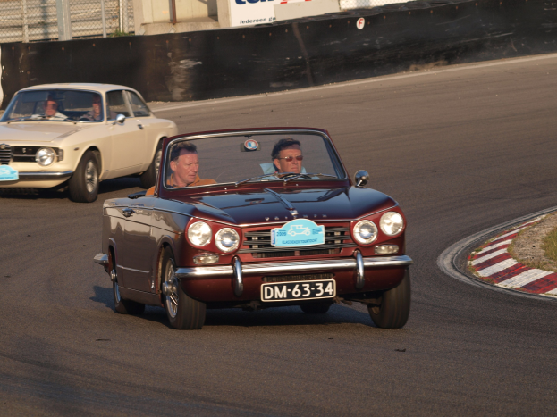 Zwei Rennautos auf einer Rennstrecke mit Zuschauern, Gras auf der rechten Seite und ein Zaun im Hintergrund mit Texttafeln.