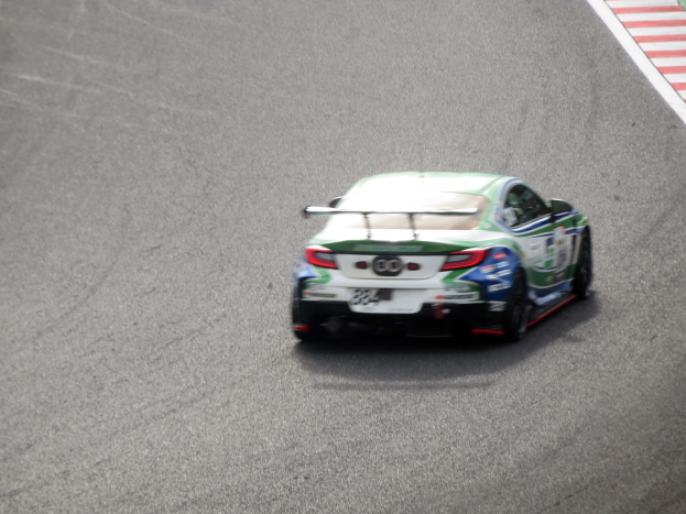 BMW M4 GT3 Rennauto auf einer Rennstrecke mit einem Rad im Vordergrund und einer Wand mit Text auf der rechten Seite.