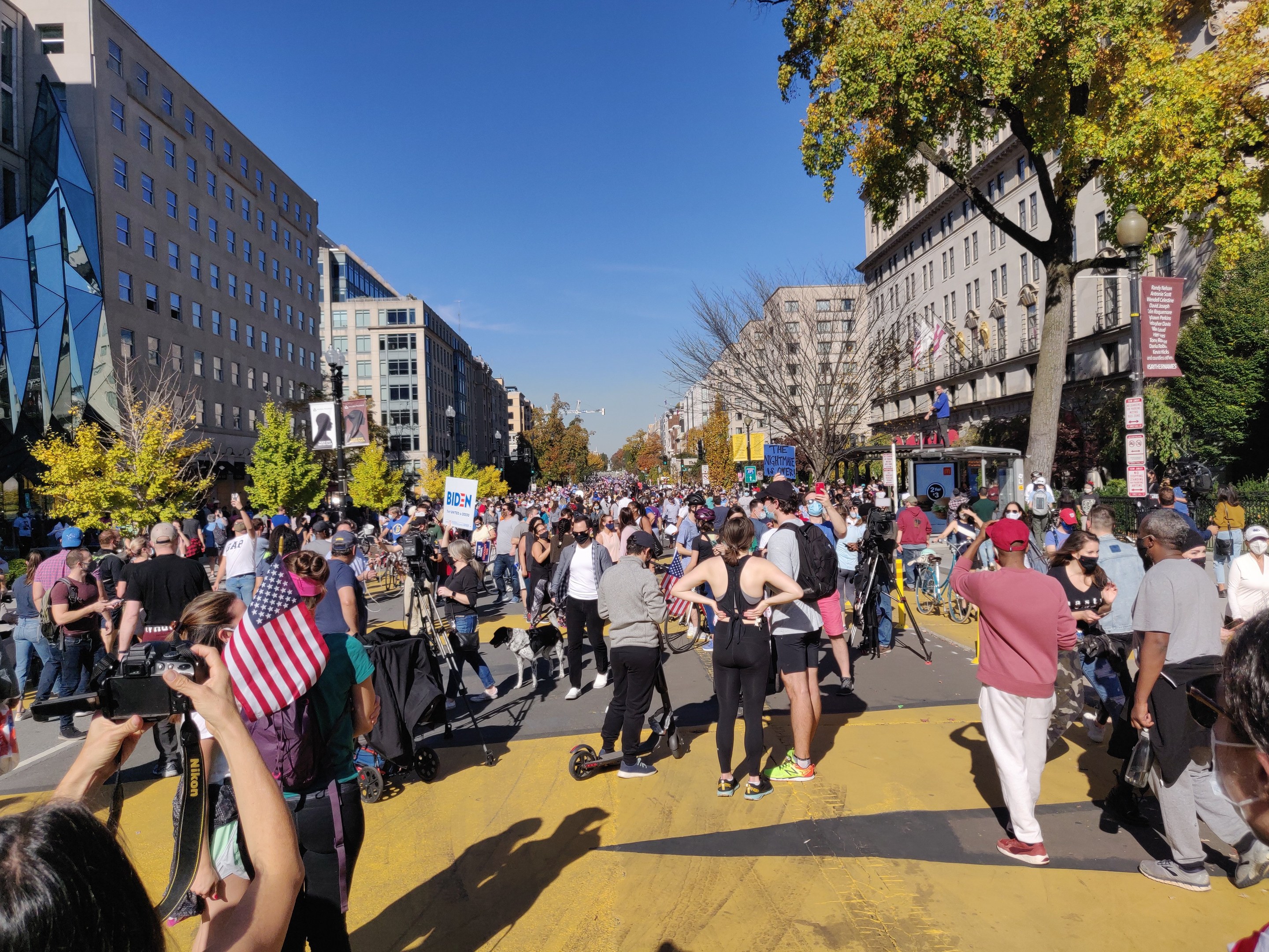Eine große Gruppe von Menschen marschiert auf einer von hohen Gebäuden gesäumten Straße mit Bäumen, Laternenmasten und einem sichtbaren Himmel, einige halten Kameras, bei einer Kundgebung in Washington, DC.