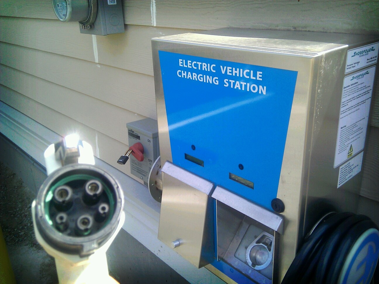 Elektrofahrzeug-Ladestation an einem Gebäude mit einem angeschlossenen Kabel und zusätzlichen an der Wand angebrachten Gegenständen im Hintergrund.