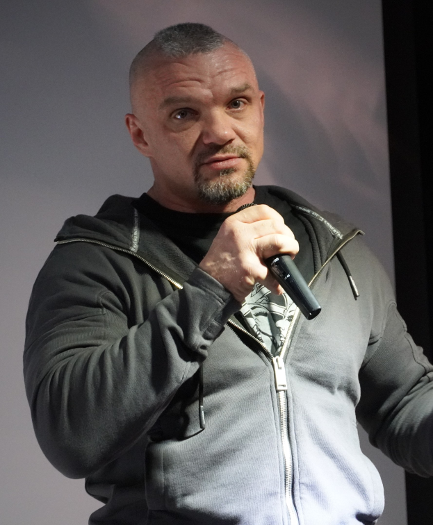 Ein Mann in einer schwarzen Kapuzenjacke und einem schwarzen Anzug mit weißem Hemd hält ein Mikrofon und steht selbstbewusst vor einer Wand.