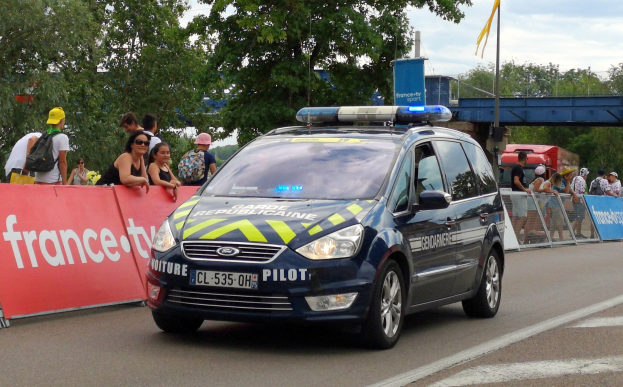 Polizeiauto fährt an einer Menschenmenge vorbei, die Schilder an Geländern hält, mit Bäumen, einer Brücke, einer Flagge und einem bewölkten Himmel im Hintergrund.