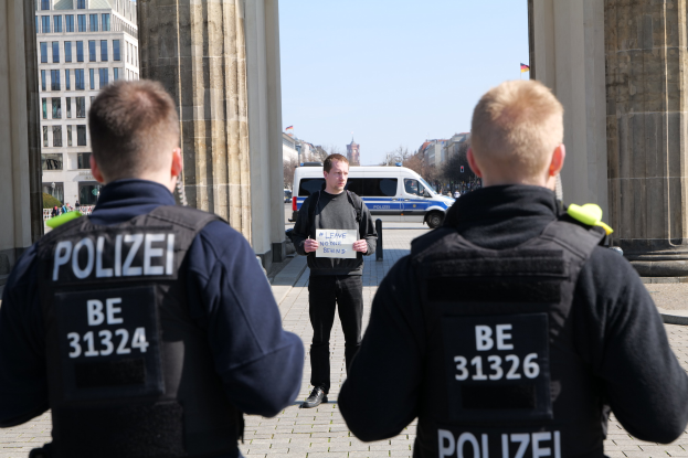 Drei Polizeibeamte stehen vor einem Gebäude, ein Mann hält ein Papier zwischen ihnen, flankiert von Säulen und einem belebten städtischen Hintergrund.