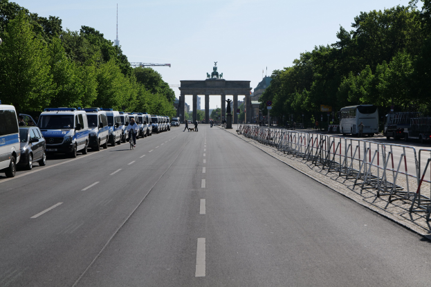 Lange Reihe von Polizeiwagen vor dem Brandenburger Tor geparkt, mit Fahrradfahrern, Passanten, Barrieren und Bäumen; ein Bogen mit Statuen im Hintergrund unter einem sichtbaren Himmel.