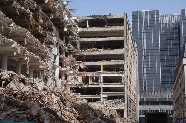Ein große Gebäude wird inmitten einer Stadt abbruch, umgeben von intakten Gebäuden, Straßeninfrastruktur, Fahrzeugen, Bäumen und einem klaren Himmel.